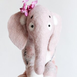 May include: A handmade pink felt elephant with white accents and a small pink flower crown. The elephant has large ears, a long trunk, and white feet. The toy is a soft, plush creation, perfect for a child's room or as a decorative item.