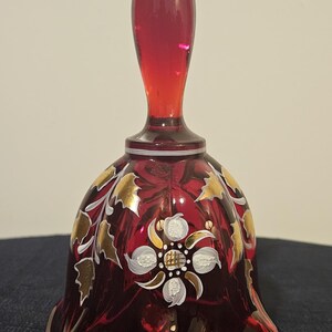 Puede incluir: Una campana decorativa de cristal rojo rubí con un mango largo y puntiagudo. La campana presenta diseños florales dorados y blancos. Este artículo es probablemente para exhibición o colección.