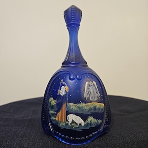 May include: A dark blue glass bell with a long handle. The bell has a painted scene of a shepherd, sheep, and an angel under a starry night sky. The bell has a decorative beaded edge.