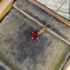 Op de afbeelding: Een gouden ketting met een ronde, gefacetteerde, dieprode edelsteen hanger in een zetting met vier pootjes. De hanger is bevestigd aan een fijne gouden touwketting die vanaf de bovenkant van het frame loopt en een klassiek sieraadontwerp toont.