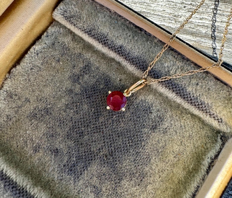 Op de afbeelding: Een gouden ketting met een ronde, gefacetteerde, dieprode edelsteen hanger in een zetting met vier pootjes. De hanger is bevestigd aan een fijne gouden touwketting die vanaf de bovenkant van het frame loopt en een klassiek sieraadontwerp toont.