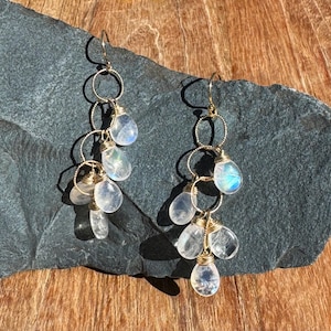 Pode incluir: Brincos de argola de ouro com múltiplas gotas de pedra da lua. Os brincos apresentam um design em cascata com múltiplos aros de ouro e contas de pedra da lua em forma de lágrima.