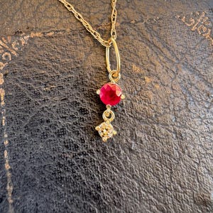 Puede incluir: Un collar de cadena dorada con un colgante que presenta una piedra preciosa roja redonda y facetada sobre una piedra transparente más pequeña de corte cuadrado. El colgante cuelga de una delicada cadena de eslabones dorados.