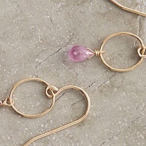 May include: A pair of gold earrings with a pink gemstone dangling from each. The earrings feature a small circle and a curved wire.