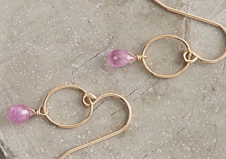 May include: A pair of gold earrings with a pink gemstone dangling from each. The earrings feature a small circle and a curved wire.