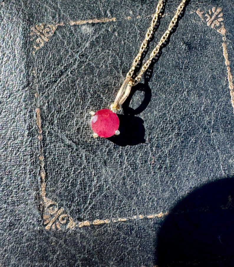 Op de afbeelding: Een gouden ketting met een ronde, gefacetteerde, levendige roze-rode edelsteen hanger in een zetting met vier pootjes. De hanger hangt aan een fijne gouden schakelketting en vertoont een helder, reflecterend oppervlak en een duidelijke steenhelderheid.
