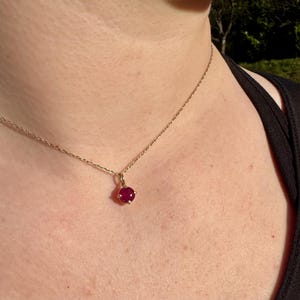 Op de afbeelding: Een fijne gouden ketting met een enkele, ronde, dieprode edelsteen hanger in een zetting met vier pootjes. De ketting rust op de huid en toont de gepolijste metalen schakels en de levendige, verzadigde kleur van de edelsteen.