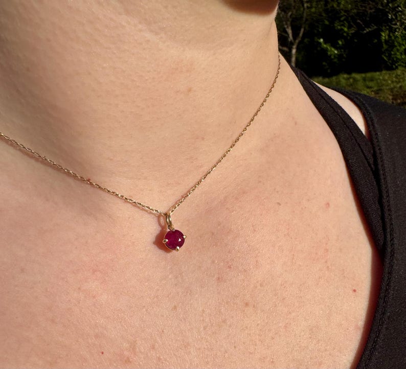 Op de afbeelding: Een fijne gouden ketting met een enkele, ronde, dieprode edelsteen hanger in een zetting met vier pootjes. De ketting rust op de huid en toont de gepolijste metalen schakels en de levendige, verzadigde kleur van de edelsteen.