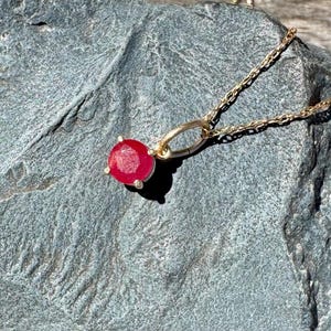 Op de afbeelding: Een gouden ketting met een enkele ronde, gefacetteerde rode edelsteen hanger in een zetting met vier pootjes. De hanger is bevestigd aan een fijne gouden schakelketting en toont een levendige karmozijnrode steen die het licht vangt.