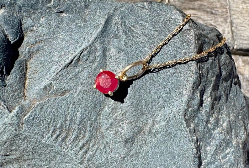 Op de afbeelding: Een gouden ketting met een enkele ronde, gefacetteerde rode edelsteen hanger in een zetting met vier pootjes. De hanger is bevestigd aan een fijne gouden schakelketting en toont een levendige karmozijnrode steen die het licht vangt.
