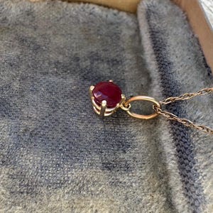 Op de afbeelding: Een gouden hanger aan een ketting met een ronde, dieprode edelsteen in een zetting met vier pootjes. De hanger zit vast aan een fijne gouden ketting met gedraaide schakels, met een gepolijste metalen afwerking en een levendige rode steen.