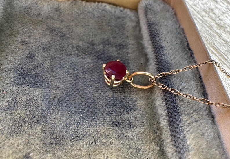Op de afbeelding: Een gouden hanger aan een ketting met een ronde, dieprode edelsteen in een zetting met vier pootjes. De hanger zit vast aan een fijne gouden ketting met gedraaide schakels, met een gepolijste metalen afwerking en een levendige rode steen.