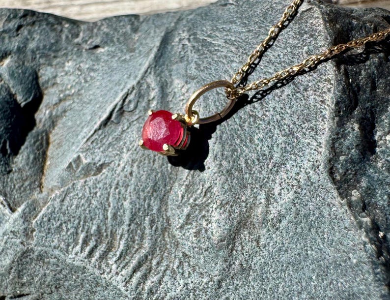 Op de afbeelding: Een gouden ketting met een enkele, ronde, gefacetteerde, felrode edelsteen hanger in een zetting met vier klauwen. De hanger hangt aan een fijne gouden schakelketting met een heldere, gepolijste metalen afwerking.