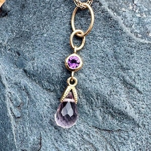 May include: A gold chain necklace featuring a pendant with a small round purple gemstone set above a larger, faceted, teardrop-shaped light purple gemstone. The pendant is suspended from a gold jump ring attached to a delicate twisted gold chain.