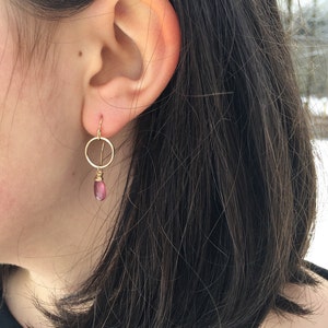 May include: Gold hoop earrings with a dangling pink gemstone. The earrings are worn in a person's ear.