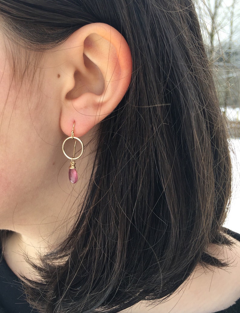 May include: Gold hoop earrings with a dangling pink gemstone. The earrings are worn in a person's ear.