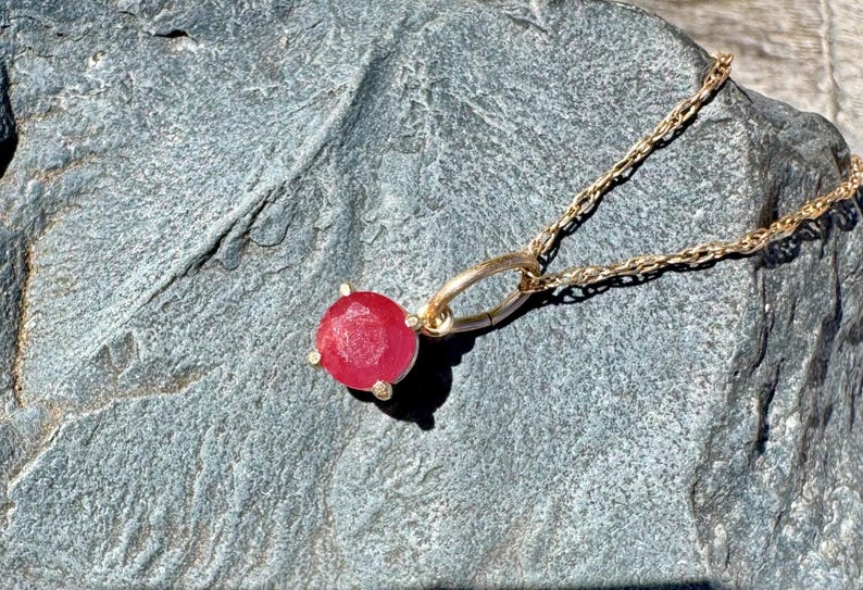 Op de afbeelding: Een gouden ketting met een ronde, gefacetteerde rode edelsteen hanger in een zetting met vier pootjes. De hanger is bevestigd aan een fijne gouden schakelketting en toont een levendige rode steen met een gestructureerde, metallic gouden afwerking.