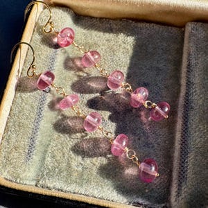 May include: Dangling earrings with pink, translucent beads and gold-colored hooks and chains. The earrings are displayed inside a vintage, velvet-lined jewelry box. The beads are a soft pink color and are strung together in a cascading design.