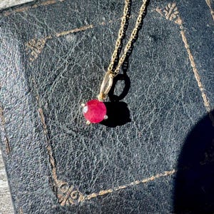 Op de afbeelding: Een gouden ketting met een enkele ronde, gefacetteerde, levendige roze-rode edelsteen hanger. De steen is bevestigd in een eenvoudige metalen zetting met kleine pootjes, hangend aan een delicate, dunne, goudkleurige schakelketting.