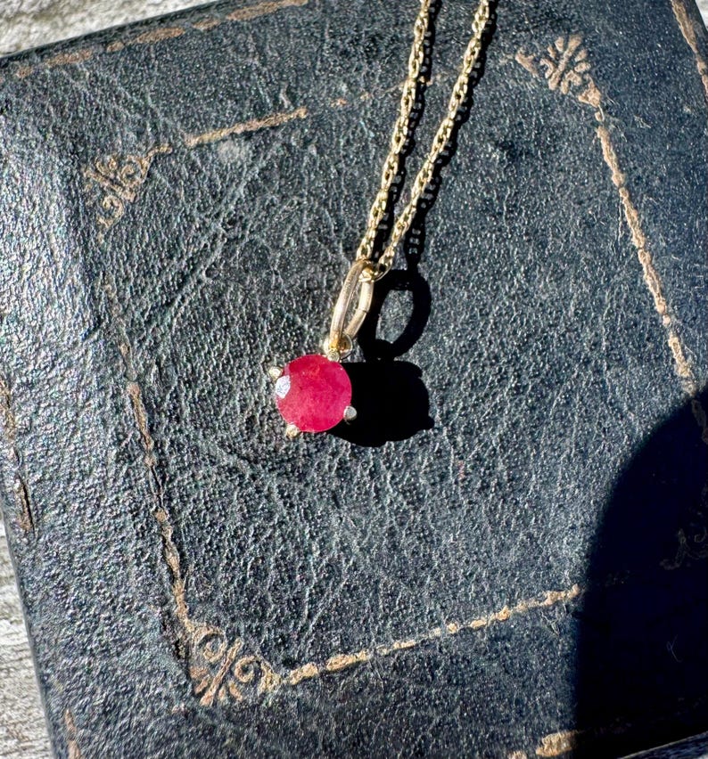 Op de afbeelding: Een gouden ketting met een enkele ronde, gefacetteerde, levendige roze-rode edelsteen hanger. De steen is bevestigd in een eenvoudige metalen zetting met kleine pootjes, hangend aan een delicate, dunne, goudkleurige schakelketting.