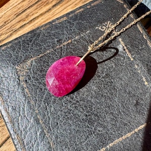 May include: A necklace featuring a faceted, teardrop-shaped, deep pink gemstone pendant suspended from a delicate gold-toned metal chain. The pendant has a textured, irregular surface and is attached to the chain by a simple gold-toned metal bail.