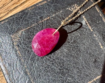 Op de afbeelding: Een ketting met een gefacetteerde, druppelvormige, dieproze edelsteen hanger aan een fijne goudkleurige metalen ketting. De hanger heeft een getextureerd, onregelmatig oppervlak en is aan de ketting bevestigd met een eenvoudig goudkleurig oogje.
