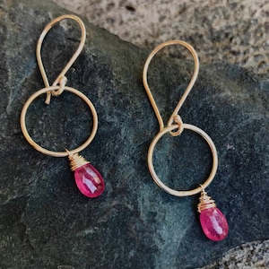 May include: Gold hoop earrings with a pink gemstone dangling from each hoop.