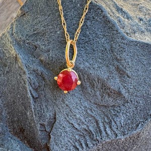 May include: A gold necklace featuring a round, faceted red gemstone pendant held by a four-prong setting. The pendant hangs from a delicate gold rope chain, showcasing a vibrant crimson stone that catches the light with its polished, multi-faceted surface.