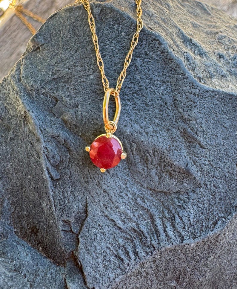 Op de afbeelding: Een gouden ketting met een ronde, gefacetteerde rode edelsteen hanger in een zetting met vier pootjes. De hanger hangt aan een fijne gouden touwketting en toont een levendige karmozijnrode steen met een gepolijst, veelzijdig oppervlak.