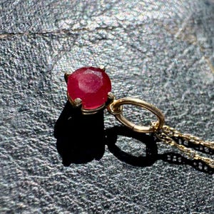 Op de afbeelding: Een hanger met een ronde, gefacetteerde, dieprode edelsteen in een vierklauws zetting van goudkleurig metaal. De hanger is met een ronde ring bevestigd aan een fijne goudkleurige ketting en heeft een gepolijste, glanzende afwerking.