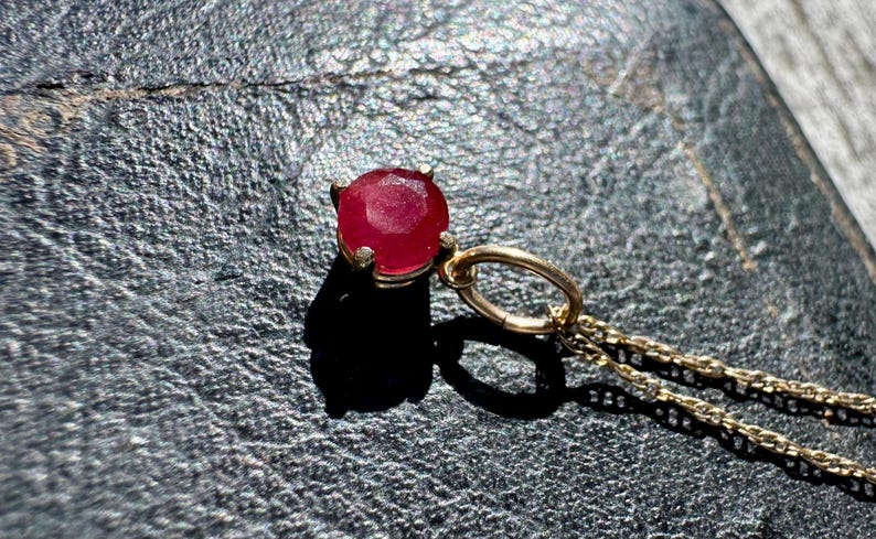 Op de afbeelding: Een hanger met een ronde, gefacetteerde, dieprode edelsteen in een vierklauws zetting van goudkleurig metaal. De hanger is met een ronde ring bevestigd aan een fijne goudkleurige ketting en heeft een gepolijste, glanzende afwerking.