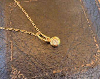 Op de afbeelding: Een gouden ketting met een enkele ronde hanger met een heldere edelsteen in een gladde zetting. De hanger is aan de ketting bevestigd met een kleine gouden ring en het metaal heeft een gepolijste afwerking die het licht weerkaatst.