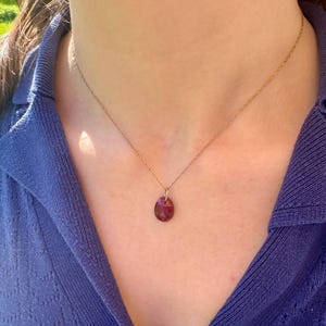 May include: A gold-colored necklace with a small, oval-shaped, dark red pendant. The pendant is suspended from a delicate chain. The necklace is worn against a blue top.