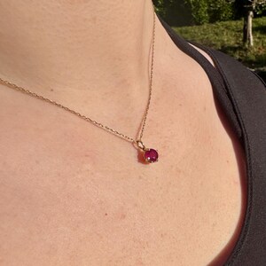 Op de afbeelding: Een fijne gouden ketting met een enkele, ronde, gefacetteerde donkerroze edelsteen als hanger, vastgezet in een zetting met vier pootjes. De ketting rust op het sleutelbeen en benadrukt de metaalglans van de ketting en de kleur van de steen.