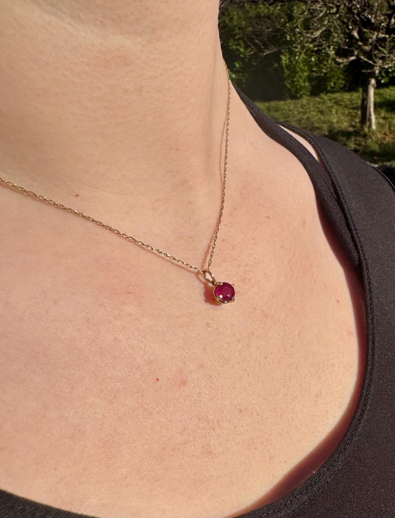 Op de afbeelding: Een fijne gouden ketting met een enkele, ronde, gefacetteerde donkerroze edelsteen als hanger, vastgezet in een zetting met vier pootjes. De ketting rust op het sleutelbeen en benadrukt de metaalglans van de ketting en de kleur van de steen.