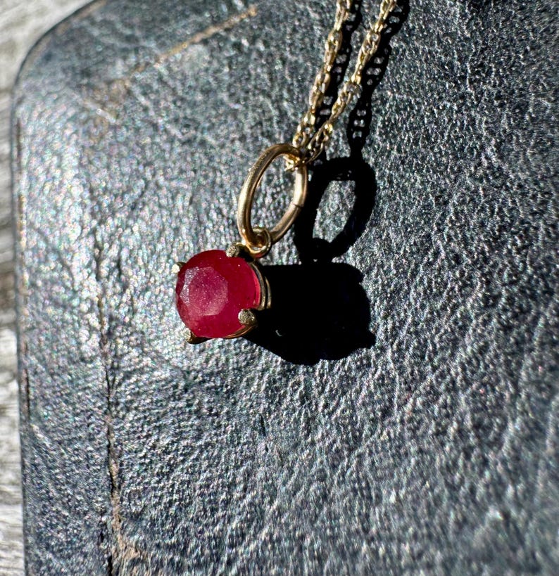 Op de afbeelding: Een gouden ketting met een ronde, gefacetteerde rode edelsteen hanger in een zetting met vier pootjes. De hanger hangt aan een fijne gouden schakelketting en toont een levendige karmozijnrode steen met een gepolijst, reflecterend oppervlak.