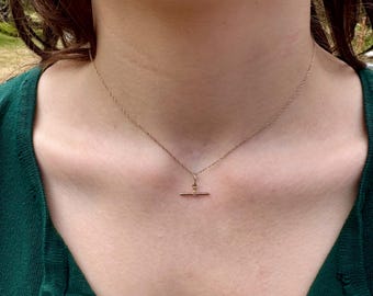 Op de afbeelding: Een delicate gouden ketting met een kleine, horizontale staafhanger die aan een centrale ring hangt. De ketting rust tegen de hals en toont een dun, gedraaid schakelontwerp dat verbonden is met de eenvoudige, rechte metalen staaf.