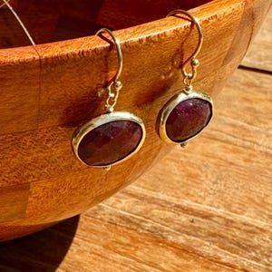 May include: A pair of gold earrings with a dark red oval gemstone. The earrings are hanging from a wooden bowl.