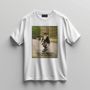 Könnte beinhalten: Weißes T-Shirt mit einer Grafik eines Mannes im Anzug, der skateboard fährt, mit dem Text "CALL ME JEWISH CUZ THE DRIP ISRAEL". Das Bild ist vor einer Straßenszene mit Gebäuden platziert.