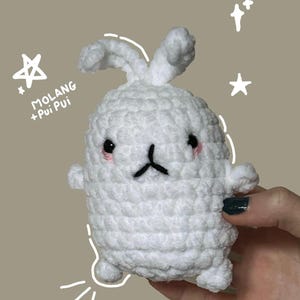 Op de afbeelding: Een handgemaakte witte gehaakte konijn knuffel met zwarte ogen, een zwarte mond en roze wangen. De knuffel heeft kleine armen en oren. De tekst "MOLANG + Pui Pui" staat aan de linkerkant van de afbeelding, met ster doodles.