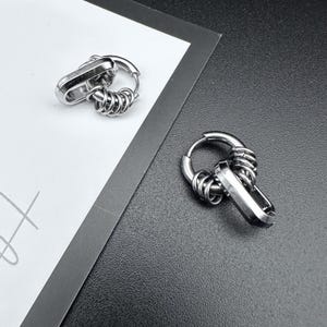 Op de afbeelding: Twee zilverkleurige oorringen. Elke oorring heeft een rechthoekige bedel bevestigd aan een spiraalvormig draadontwerp, verbonden met een cirkelvormige ring. De oorbellen worden gepresenteerd op een wit en zwart oppervlak.