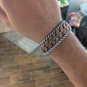 May include: A copper and silver-toned bracelet with a chain-link design. The bracelet is worn on a wrist and features a braided silver border. The copper links are interwoven with silver links.