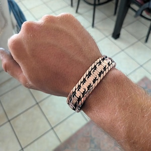 May include: A copper-colored cuff bracelet featuring a central row of repeating, wave-like patterns flanked by two twisted rope-style borders. The metal has a polished finish with dark, recessed areas that highlight the intricate texture of the design.