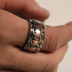 May include: A silver-toned ring with a woven design and copper-colored accents. The ring is on a finger, showcasing the intricate pattern and metallic contrast. The jewelry piece has a bold, textured appearance.