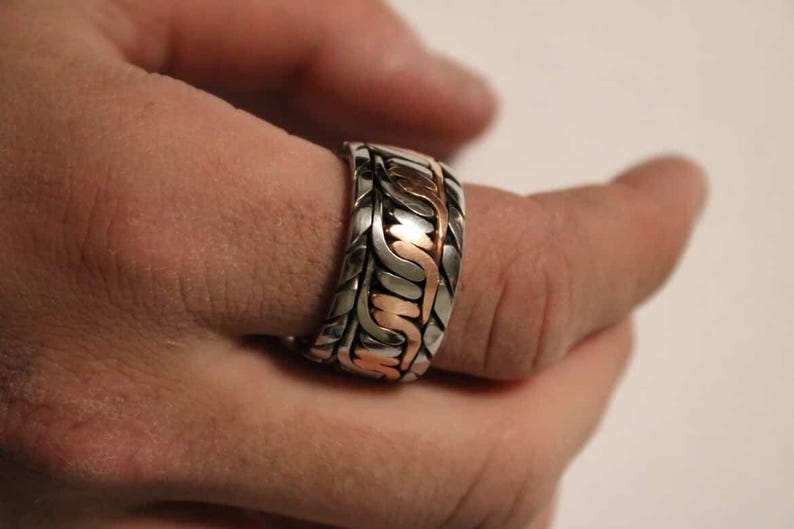 May include: A silver-toned ring with a woven design and copper-colored accents. The ring is on a finger, showcasing the intricate pattern and metallic contrast. The jewelry piece has a bold, textured appearance.