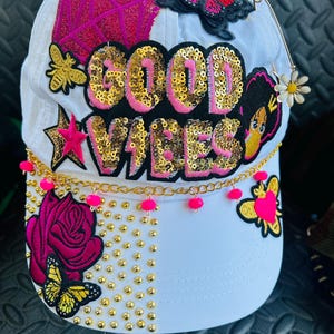 May include: White baseball cap with sequined "GOOD VIBES" text, pink accents, and gold embellishments. Features butterfly and bee appliqués, a pink star, and a rose. A gold chain with pink beads adorns the brim.