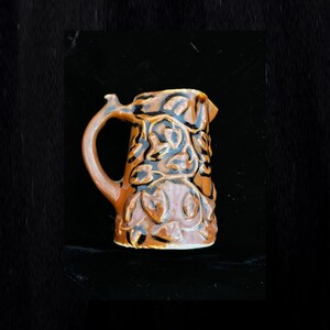 May include: A brown ceramic pitcher with a black floral design. The pitcher has a handle and a spout.