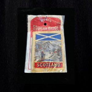 May include: A gold-colored souvenir badge with a blue and white Scottish flag, a skier on a snowy mountain, and the word "SCOTLAND" in red letters.