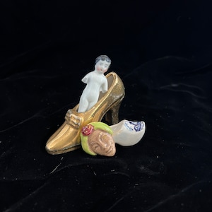 May include: A porcelain figurine of a person standing in a gold shoe. The shoe has a black heel and a white and blue clog is lying next to it.