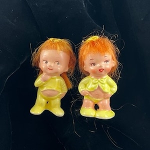 May include: Two ceramic dolls with yellow dresses and orange hair. The dolls have painted facial features and are wearing yellow shoes.
